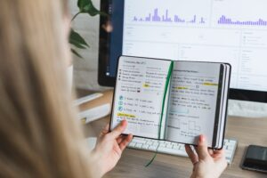 Woman reading notes from notebook in front of her computer screen.